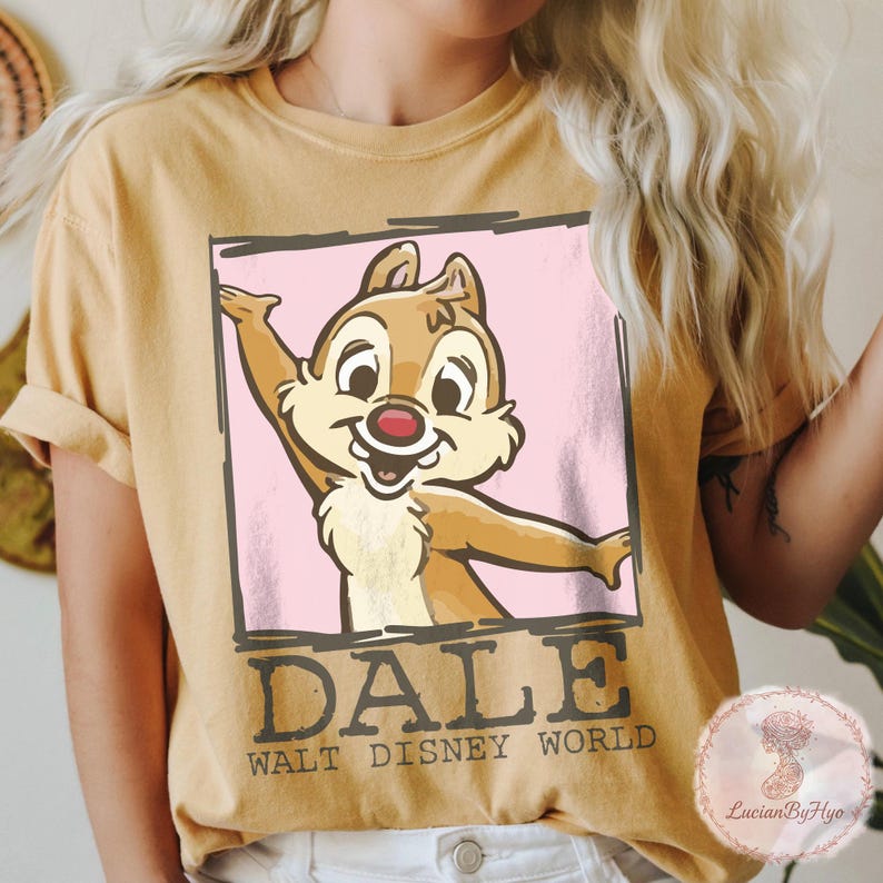 Puede incluir: Camiseta de color mostaza con una imagen de dibujos animados de Dale de Chip y Chop, los guardianes rescatadores de Disney. Dale se representa con una expresi&oacute;n alegre, enmarcado en un recuadro rosa. El texto "DALE WALT DISNEY WORLD" est&aacute; impreso debajo.