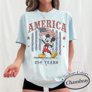 Pu&ograve; includere: T-shirt azzurra con una grafica invecchiata di Mickey Mouse con una bandiera americana. La parola "AMERICA" &egrave; sopra l'immagine, con "SINCE 1776 - 250 YEARS" sotto. La maglietta &egrave; etichettata "Comfort Colors Chambray".