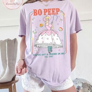 May include: A lilac-colored t-shirt featuring a cartoon illustration of Bo Peep from Toy Story. The design includes the text "BO PEEP" and "YOU'VE GOT A FRIEND IN ME Est. 1997". The graphic is framed with a colorful border.
