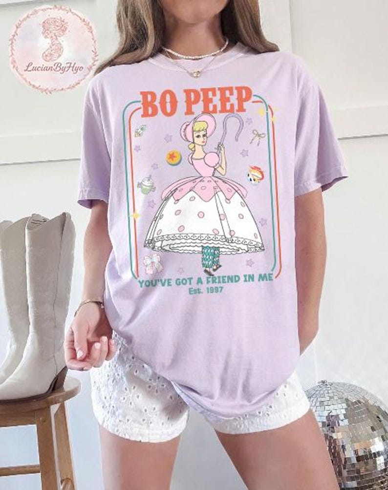 May include: A lilac-colored t-shirt featuring a cartoon illustration of Bo Peep from Toy Story. The design includes the text "BO PEEP" and "YOU'VE GOT A FRIEND IN ME Est. 1997". The graphic is framed with a colorful border.