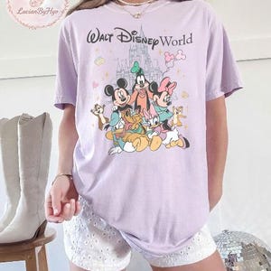 May include: A light purple t-shirt featuring a graphic of Disney characters, including Mickey Mouse, Minnie Mouse, Goofy, and Donald Duck, with the text "Walt Disney World". The design includes a castle and other cartoon elements.