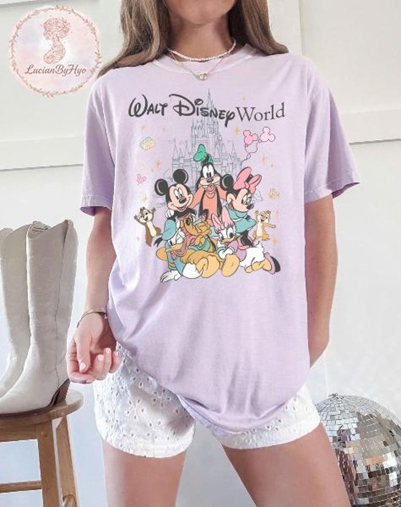 May include: A light purple t-shirt featuring a graphic of Disney characters, including Mickey Mouse, Minnie Mouse, Goofy, and Donald Duck, with the text "Walt Disney World". The design includes a castle and other cartoon elements.