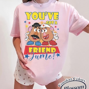 Op de afbeelding: Lichtroze T-shirt met een afbeelding van twee aardappelvormige personages en de tekst "YOU'VE GOT A FRIEND Jume!". Het ontwerp bevat blauwe, gele en rode accenten. Het shirt is gemarkeerd met "Comfort Colors Blossom".