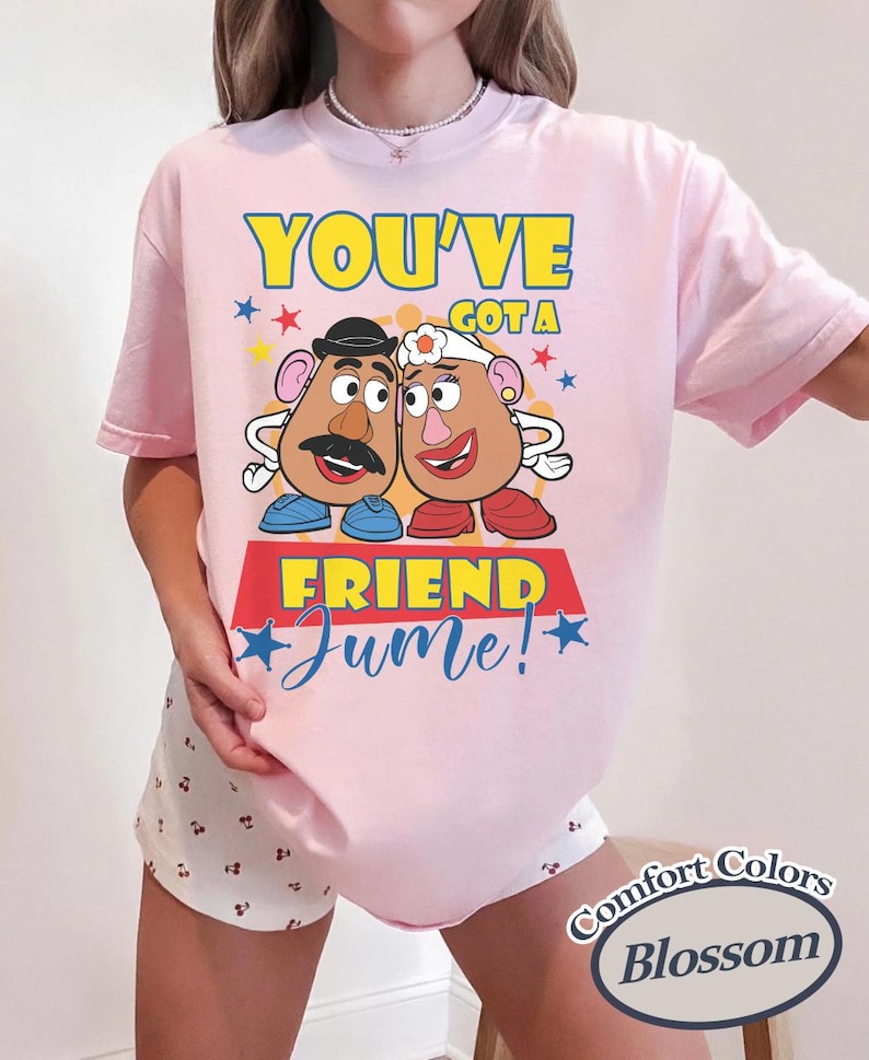 Op de afbeelding: Lichtroze T-shirt met een afbeelding van twee aardappelvormige personages en de tekst "YOU'VE GOT A FRIEND Jume!". Het ontwerp bevat blauwe, gele en rode accenten. Het shirt is gemarkeerd met "Comfort Colors Blossom".