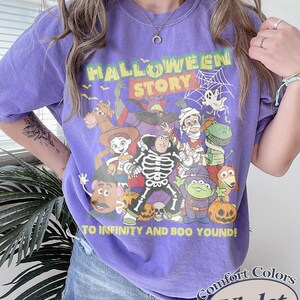May include: Violet Comfort Colors t-shirt with a Halloween Story graphic featuring cartoon characters. The design includes the text "TO INFINITY AND BOO YOUND!" and a spiderweb. The shirt is a crew neck.