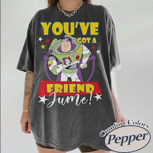 Op de afbeelding: Donkergrijs T-shirt met Buzz Lightyear uit Toy Story en de tekst "YOU'VE GOT A FRIEND June!". Het ontwerp bevat gele, groene, paarse en rode accenten. Het shirt heeft een vintage, gewassen look.