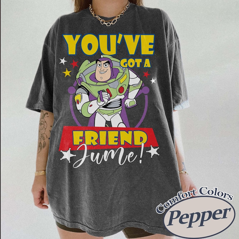 Op de afbeelding: Donkergrijs T-shirt met Buzz Lightyear uit Toy Story en de tekst "YOU'VE GOT A FRIEND June!". Het ontwerp bevat gele, groene, paarse en rode accenten. Het shirt heeft een vintage, gewassen look.