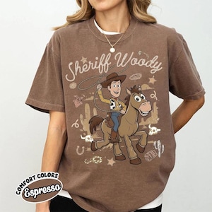 May include: A brown short-sleeved t-shirt featuring a graphic print of the character Sheriff Woody riding a horse. The design includes a lasso, cacti, stars, and the text Sheriff Woody in a cursive font. The shirt has a crew neckline and a relaxed fit.