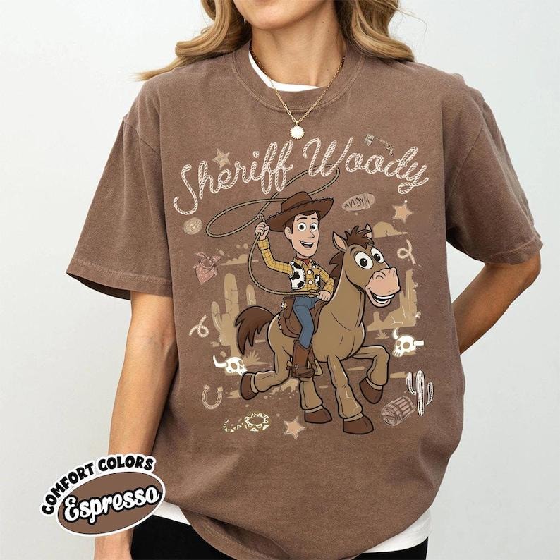 May include: A brown short-sleeved t-shirt featuring a graphic print of the character Sheriff Woody riding a horse. The design includes a lasso, cacti, stars, and the text Sheriff Woody in a cursive font. The shirt has a crew neckline and a relaxed fit.