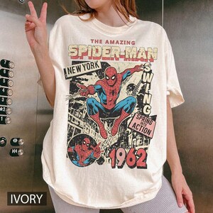 May include: An ivory-colored t-shirt featuring a graphic print of Spider-Man in a dynamic pose. The text reads THE AMAZING SPIDER-MAN, NEW YORK, SWING, SPRING INTO ACTION, and 1962. The design includes a comic book style illustration of the character.