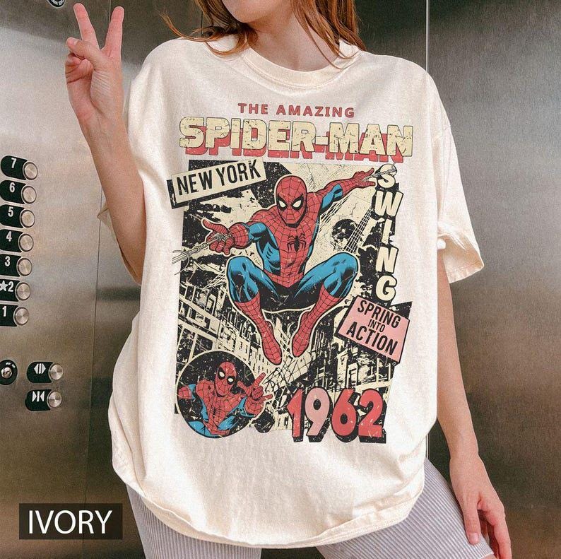 May include: An ivory-colored t-shirt featuring a graphic print of Spider-Man in a dynamic pose. The text reads THE AMAZING SPIDER-MAN, NEW YORK, SWING, SPRING INTO ACTION, and 1962. The design includes a comic book style illustration of the character.