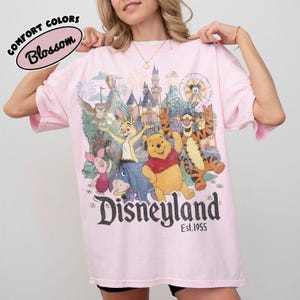May include: A light pink t-shirt featuring a graphic print of Winnie the Pooh, Piglet, Tigger, Eeyore, and Rabbit in front of a castle. The text reads Disneyland Est. 1955. A small oval label in the top left corner says Comfort Colors Blossom.