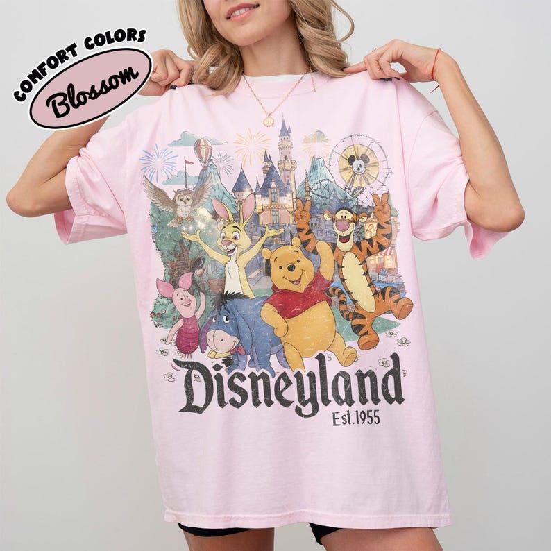 May include: A light pink t-shirt featuring a graphic print of Winnie the Pooh, Piglet, Tigger, Eeyore, and Rabbit in front of a castle. The text reads Disneyland Est. 1955. A small oval label in the top left corner says Comfort Colors Blossom.