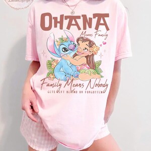May include: Pink t-shirt featuring a graphic of Stitch and Lilo from Lilo & Stitch, with the words "Ohana Means Family" and "Family Means Nobody Gets Left Behind or Forgotten". The design includes floral accents and hearts.