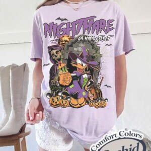 May include: Orchid-colored t-shirt with a Halloween-themed graphic. The design features a cartoon duck in a witch hat, pumpkins, and the text "Nightmare on Main Street." Bats and a tombstone complete the spooky scene.