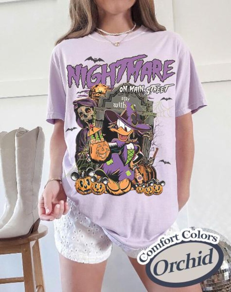 May include: Orchid-colored t-shirt with a Halloween-themed graphic. The design features a cartoon duck in a witch hat, pumpkins, and the text "Nightmare on Main Street." Bats and a tombstone complete the spooky scene.