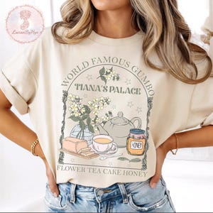 May include: Beige t-shirt with a graphic featuring the text "WORLD FAMOUS GUMBO" and "TIANA'S PALACE". The design includes a teapot, flowers, honey, cake, and tea. The words "FLOWER TEA CAKE HONEY" are also printed on the shirt.
