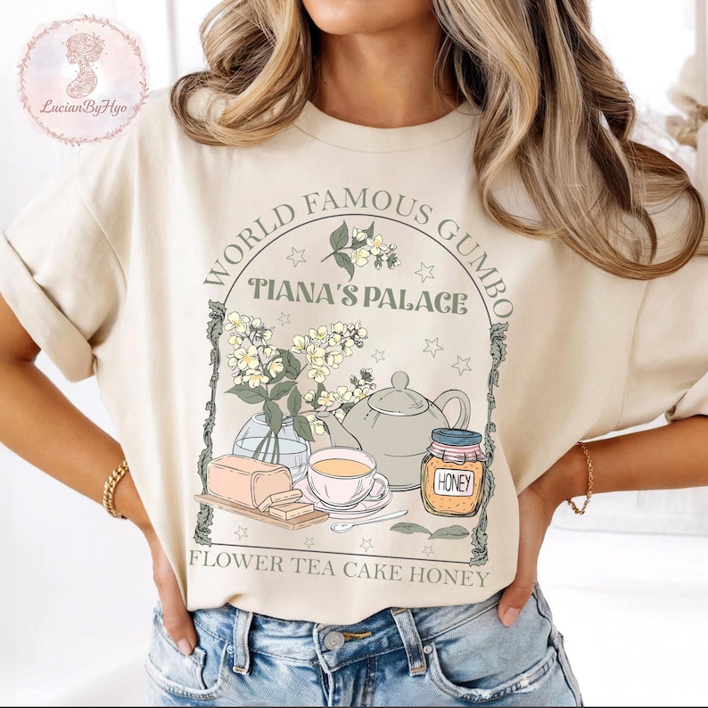 May include: Beige t-shirt with a graphic featuring the text "WORLD FAMOUS GUMBO" and "TIANA'S PALACE". The design includes a teapot, flowers, honey, cake, and tea. The words "FLOWER TEA CAKE HONEY" are also printed on the shirt.