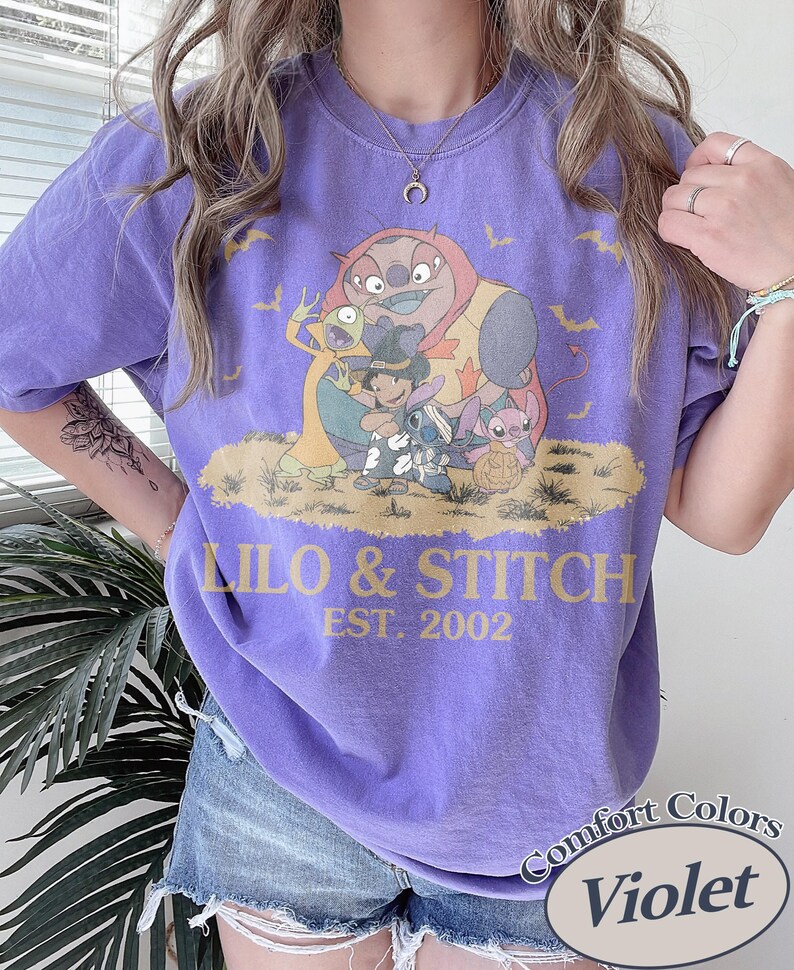 K&ouml;nnte beinhalten: Violettes T-Shirt mit einem Aufdruck von Lilo & Stitch-Charakteren. Das Design enth&auml;lt den Text "LILO & STITCH EST. 2002" in goldfarbenen Buchstaben. Das Shirt hat auch ein "Comfort Colors"-Etikett mit dem Wort "Violet".
