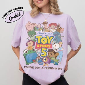 May include: A light purple t-shirt featuring a graphic print of various Toy Story characters, the text Disney Pixar Toy Story 5, and the phrase You've Got A Friend In Me. A small oval label in the top left corner reads Comfort Colors Orchid.