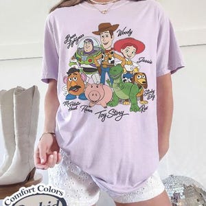 May include: A lilac Comfort Colors t-shirt with a graphic of Toy Story characters, including Woody, Buzz Lightyear, and Jessie. The shirt has the words "Toy Story" printed below the characters. The shirt is labelled "Orchid".
