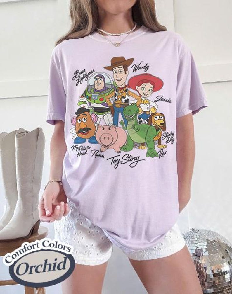 May include: A lilac Comfort Colors t-shirt with a graphic of Toy Story characters, including Woody, Buzz Lightyear, and Jessie. The shirt has the words "Toy Story" printed below the characters. The shirt is labelled "Orchid".