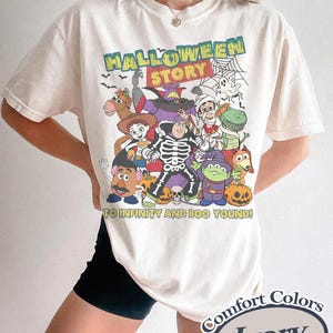 May include: Ivory-colored t-shirt featuring a Halloween-themed graphic with characters from a popular animated movie. The design includes the text "HALLOWEEN STORY" and "TO INFINITY AND BOO YOUND!" in a playful font. The shirt is labeled "Comfort Colors".