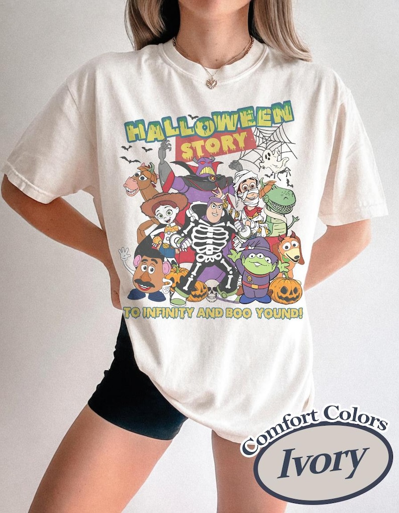 May include: Ivory-colored t-shirt featuring a Halloween-themed graphic with characters from a popular animated movie. The design includes the text "HALLOWEEN STORY" and "TO INFINITY AND BOO YOUND!" in a playful font. The shirt is labeled "Comfort Colors".