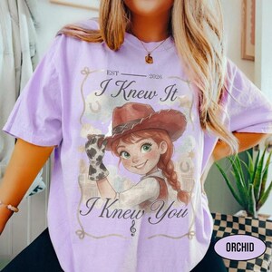 May include: A light purple t-shirt featuring a graphic of a cartoon character wearing a brown cowboy hat and a cow-print glove. The text reads "EST 2026", "I Knew It", and "I Knew You" in a decorative font, framed by a rope-like border design.