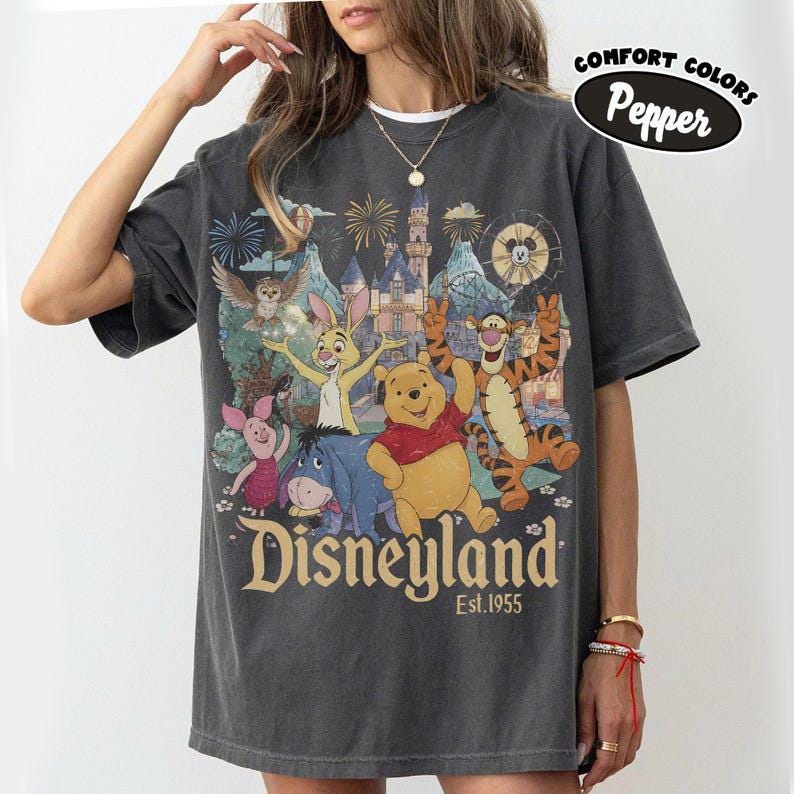 May include: A charcoal grey short-sleeve t-shirt featuring a graphic print of Winnie the Pooh, Tigger, Piglet, Eeyore, and Rabbit in front of a castle. The text reads Disneyland Est. 1955. A circular badge in the corner says Comfort Colors Pepper.