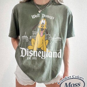 Puede incluir: Camiseta verde oliva con un dise&ntilde;o de Pluto de Disney, el logo de Disneyland y el texto "Est. 1955". La camiseta est&aacute; etiquetada como "Comfort Colors Moss."