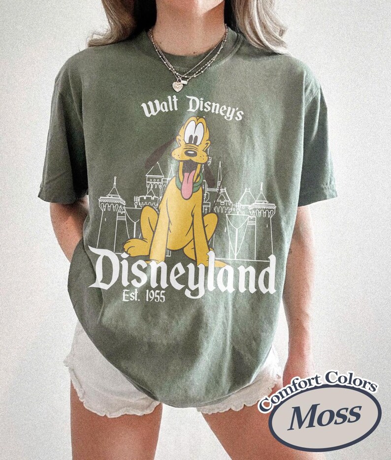 Puede incluir: Camiseta verde oliva con un dise&ntilde;o de Pluto de Disney, el logo de Disneyland y el texto "Est. 1955". La camiseta est&aacute; etiquetada como "Comfort Colors Moss."
