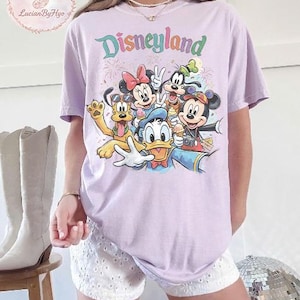 May include: A light purple t-shirt featuring the word "Disneyland" in a colorful, whimsical font. The shirt showcases a graphic of Disney characters, including Mickey Mouse, Minnie Mouse, Donald Duck, Goofy, and Pluto. The design includes fireworks and ice cream.