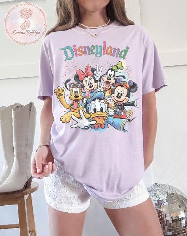 May include: A light purple t-shirt featuring the word "Disneyland" in a colorful, whimsical font. The shirt showcases a graphic of Disney characters, including Mickey Mouse, Minnie Mouse, Donald Duck, Goofy, and Pluto. The design includes fireworks and ice cream.