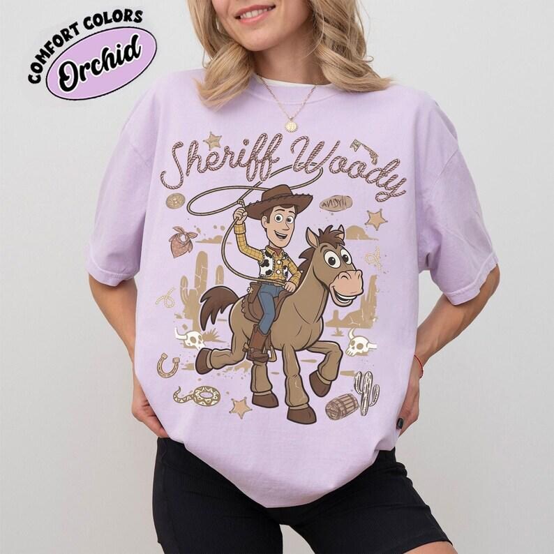 May include: A light purple t-shirt featuring a graphic of Sheriff Woody riding a brown horse. The text "Sheriff Woody" is printed above the characters. Additional western-themed icons include cacti, stars, a horseshoe, and a snake.