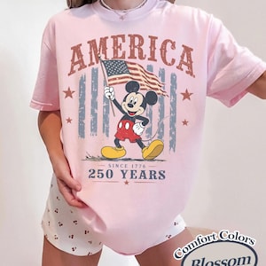 Pu&ograve; includere: T-shirt rosa chiaro con una grafica di Mickey Mouse che tiene una bandiera americana. La parola "AMERICA" &egrave; stampata sopra Mickey, con "SINCE 1776" e "250 YEARS" sotto. La maglietta ha un'etichetta "Comfort Colors Blossom".