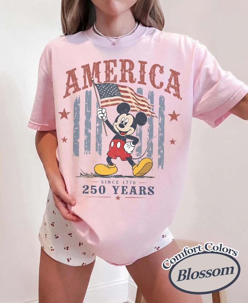Pu&ograve; includere: T-shirt rosa chiaro con una grafica di Mickey Mouse che tiene una bandiera americana. La parola "AMERICA" &egrave; stampata sopra Mickey, con "SINCE 1776" e "250 YEARS" sotto. La maglietta ha un'etichetta "Comfort Colors Blossom".