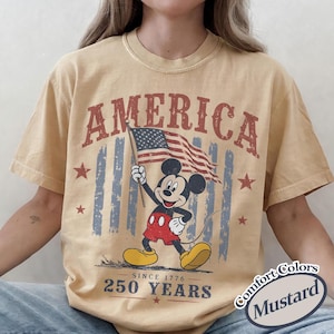 Pu&ograve; includere: T-shirt color senape con grafica vintage di Mickey Mouse con bandiera americana. La scritta "AMERICA" &egrave; sopra il personaggio, con "250 YEARS SINCE 1776" sotto. La maglietta ha un aspetto vintage.