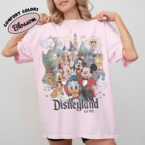 Peut inclure: Un t-shirt rose clair avec une impression graphique de Mickey Mouse, Minnie Mouse, Donald Duck, Dingo et Pluto devant un ch&acirc;teau et une grande roue. Le texte indique Disneyland Est. 1955. Une &eacute;tiquette Comfort Colors Blossom est visible.