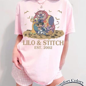 K&ouml;nnte beinhalten: Hellrosa T-Shirt mit einer Grafik von Lilo & Stitch-Charakteren und dem Text "LILO & STITCH EST. 2002". Das Shirt ist ein Comfort Colors Blossom T-Shirt.