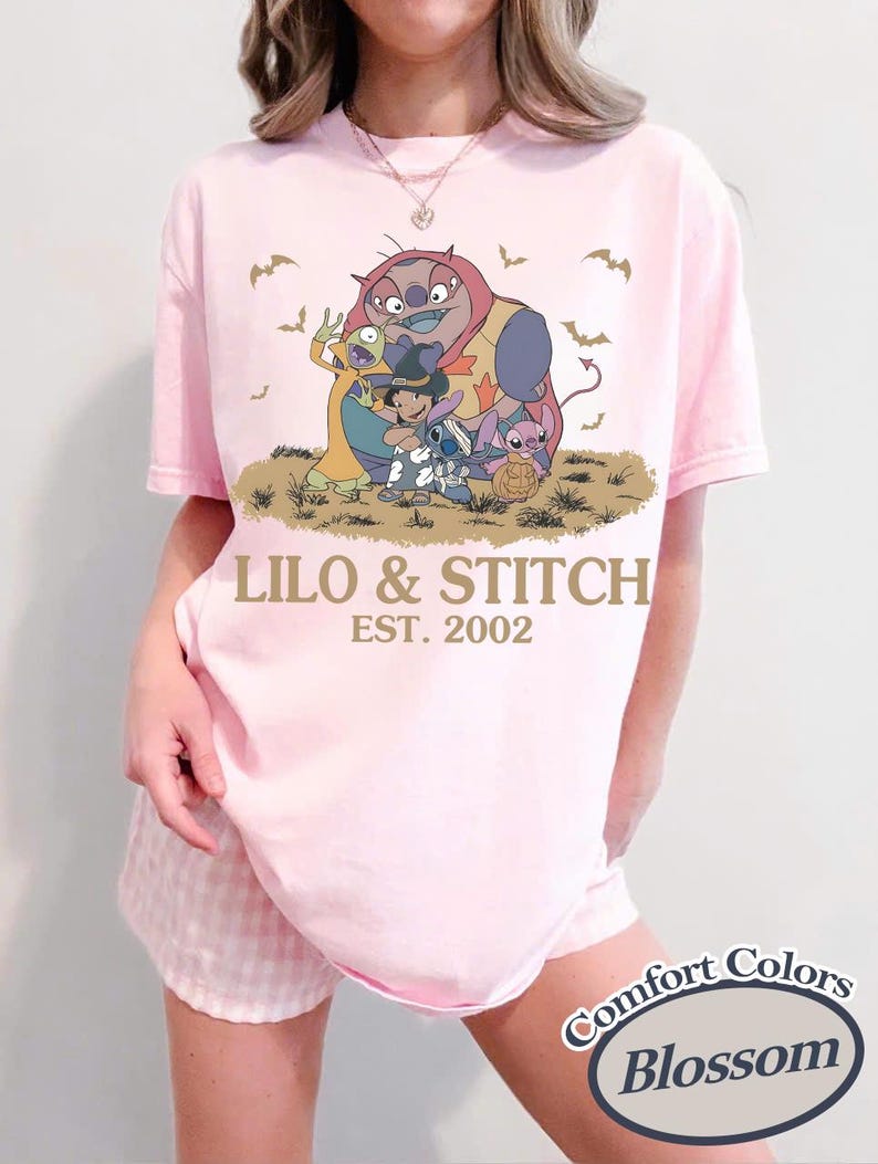 K&ouml;nnte beinhalten: Hellrosa T-Shirt mit einer Grafik von Lilo & Stitch-Charakteren und dem Text "LILO & STITCH EST. 2002". Das Shirt ist ein Comfort Colors Blossom T-Shirt.