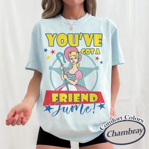 Op de afbeelding: Lichtblauw T-shirt met een cartoon illustratie van een personage met een herdersstaf, met de tekst "YOU'VE GOT A FRIEND Jume!" in geel en blauw. Het shirt heeft een ronde hals en korte mouwen.