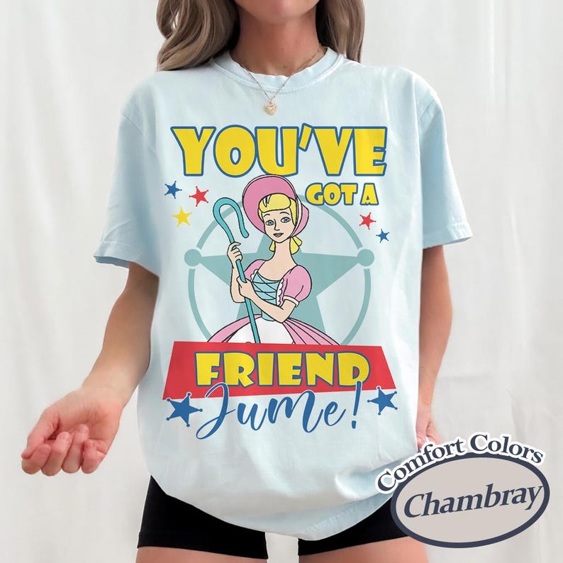 Op de afbeelding: Lichtblauw T-shirt met een cartoon illustratie van een personage met een herdersstaf, met de tekst "YOU'VE GOT A FRIEND Jume!" in geel en blauw. Het shirt heeft een ronde hals en korte mouwen.