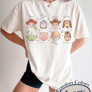 May include: Ivory-colored t-shirt featuring cartoon illustrations of characters from a popular animated movie. The shirt includes images of Jessie, Buzz Lightyear, Woody, Slinky, Alien, Hamm, Bo Peep, and Bullseye. The text "Comfort Colors" and "Ivory" are also visible.