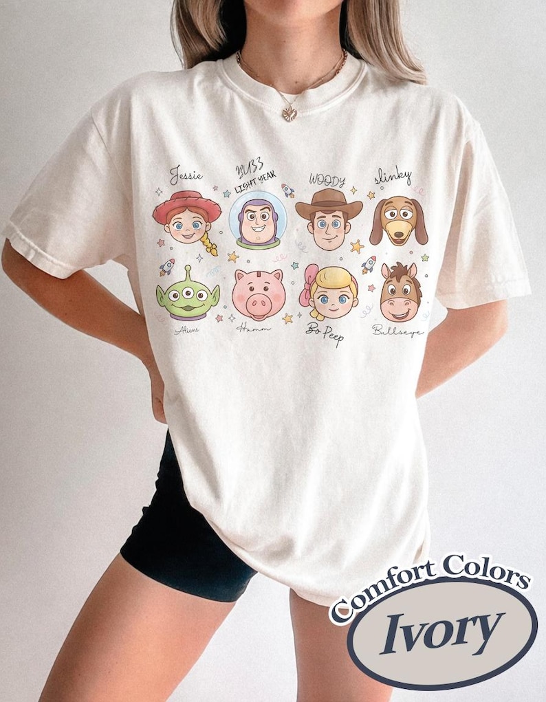 May include: Ivory-colored t-shirt featuring cartoon illustrations of characters from a popular animated movie. The shirt includes images of Jessie, Buzz Lightyear, Woody, Slinky, Alien, Hamm, Bo Peep, and Bullseye. The text "Comfort Colors" and "Ivory" are also visible.