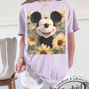Puede incluir: Camiseta lila claro con un dise&ntilde;o de Mickey Mouse sonriente rodeado de girasoles. La camiseta tiene mangas cortas y un corte holgado. El texto "Comfort Colors Orchid" est&aacute; impreso en una pegatina en la esquina inferior derecha.