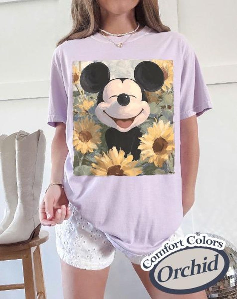 Puede incluir: Camiseta lila claro con un dise&ntilde;o de Mickey Mouse sonriente rodeado de girasoles. La camiseta tiene mangas cortas y un corte holgado. El texto "Comfort Colors Orchid" est&aacute; impreso en una pegatina en la esquina inferior derecha.
