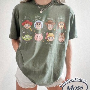 May include: Moss green t-shirt featuring cartoon character portraits. The shirt includes characters like Jessie, Buzz Lightyear, Woody, and Slinky. The text "Comfort Colors" and "Moss" are printed on the bottom right.