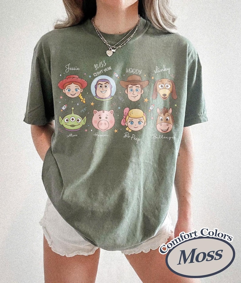 May include: Moss green t-shirt featuring cartoon character portraits. The shirt includes characters like Jessie, Buzz Lightyear, Woody, and Slinky. The text "Comfort Colors" and "Moss" are printed on the bottom right.