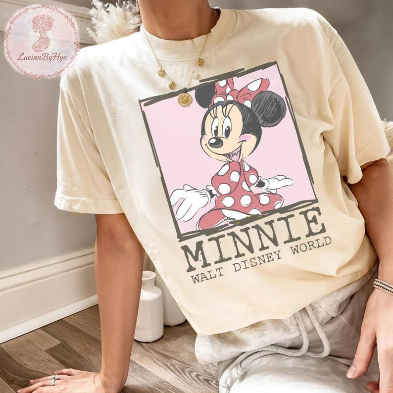 Puede incluir: Camiseta color crema con un gr&aacute;fico de Minnie Mouse en un marco rosa. Minnie lleva un vestido y un lazo de lunares rojos. El texto "MINNIE WALT DISNEY WORLD" est&aacute; impreso debajo.